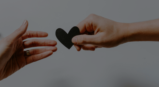 A photograph of a hand reaching across the frame, handing a paper cut-out of a heart to another hand.